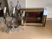 View of metal chandelier with ornate design, white candle sleeves, and hanging glass crystals, alongside wooden shadow box with vintage metal hardware.