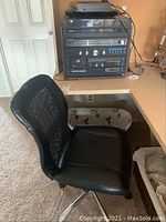 Side view of black office chair with mesh back, showing padded seat and chrome base with wheels. Stereo system visible in background.
