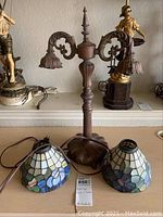 Photo showing lamp base with two ornate curved arms and two stained glass style shades placed in front.