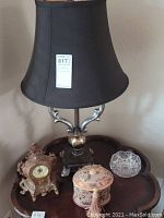 Photo showing lamp with metallic base and black shade, ornate clock, fabric box, and glass bowl on wooden table