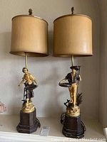 Pair of vintage figurine table lamps with beige lampshades on wooden bases, shown side by side