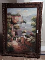 Full view of oversized framed artwork depicting a scenic coastal village by water with a path and greenery.