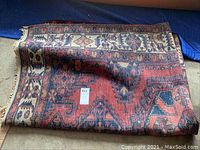 Folded Persian rug partially rolled on concrete floor showing overall pattern and wear