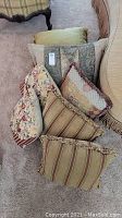Seven decorative pillows arranged on a carpet near furniture, showing variety of patterns and sizes.
