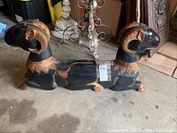 Photo of full double ram headed statue showing black wood body with gold and red painted horns and collars, placed on concrete floor with tag attached.