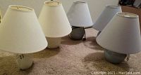 Photo showing five ceramic table lamps with white tapered lampshades placed on carpeted floor. Bases are in blue and white ceramic with some decorative designs visible.