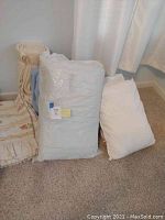 Various bed pillows, comforter in package, blanket, and cylindrical pillows against wall with curtain.
