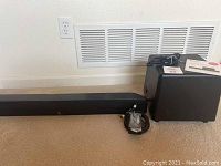 Full view showing black Boston Acoustics soundbar, black wireless subwoofer standing upright, power cable, and remote control.