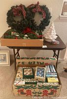 Photo showing three green Christmas wreaths with red plaid bows on a table with a white ceramic Christmas tree beside them and below, a large box of Christmas ornaments.