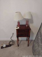 Wood end table with two drawers, vintage cranberry glass lamps on top, against wall with visible cords and power strips on floor