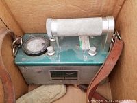 Metal box Geiger counter with green top, analog meter, control knobs, and leather strap inside a cardboard box.