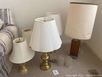 Five table lamps grouped on floor with corded tall amber glass lamp having ornate metal base and cream oval lampshade. Four other lamps smaller with white or cream shades and brass bases. Two clear ribbed glass pendant shades visible on floor.