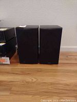 Pair of black bookshelf speakers standing side by side on a wooden floor, fronts covered with fabric grilles.