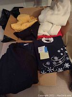 Two boxes filled with folded women's tops and sweaters in various colors including cream, yellow, black, navy blue with snowflake design, and red.