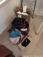 Multiple bags including a large brown paper bag filled with bras and intimates and a green bag containing assorted clothing items and socks, as well as a box with slippers.