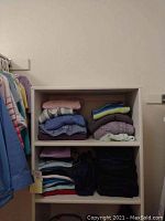 Folded sweaters, knit tops, and jeans stacked on two shelves in white shelving unit