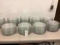 View of multiple stacks of clear, round glass plates with embossed rings on rim, set on a white table.