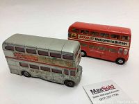 Photo showing two vintage die cast metal Corgi British double decker bus toys, one red and one silver, both around 5 inches long, placed side by side on a white background with a MaxSold card for scale.