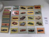 Full view of 25 trading cards spread out in rows with remaining cards stacked to the left. Cards in protective hard plastic sleeves showing multiple different colored backgrounds and vintage cars illustrated.