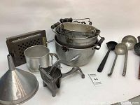 View of all 13 items including camping pots, ladles, funnel, cup, popcorn maker, cast iron ladle and metal grate.