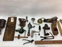 Photo showing a variety of antique wooden and metal hand tools arranged on a white surface including planes, drills, clamps, and measuring devices.