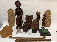 Collection of 13 wooden and stone carvings including tall drummer figurine, carved panels, masks, small sculptures and flutes arranged on white surface.