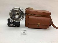 Full view of Kodak Brownie camera with attached large silver flash beside brown leather carrying case with shoulder strap.