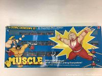 Front of original blue cardboard box for M.U.S.C.L.E. rubber wrestler toy set. Box opened showing many small rubber wrestler figures in plastic trays lined behind a plastic window. Colorful cartoon wrestler art and logos printed on box front.