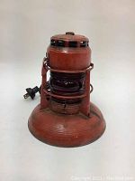 Full front view of the vintage metal lantern showing its orange-red paint, rust spots, electric cord and plug, glass shade, and hanging handle.
