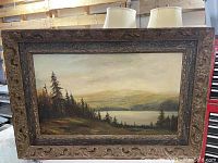 Full view of framed oil painting depicting a landscape with pine trees and lake.