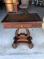 Front view showing the entire antique harp table with ornate harp-shaped carved wooden base, claw feet, drawer with brass knob, and rectangular leather-topped surface.