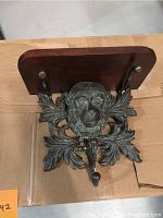 Front view showing brass monkey face with detailed foliage design and three hat rack hooks below.