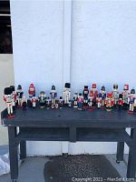 Wide view showing a collection of 21 wooden Christmas nutcracker figurines of various sizes and conditions arranged on a black table against an outdoor wall.