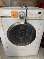 Front view of the Frigidaire washing machine showing the circular glass door, power cord draped over the top, and orange lot number sticker.