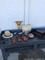 Photo of all items on a black table including hat, violins, figurine, picture frame, wall sconce, trinket box, and wooden box