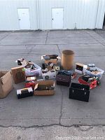 Wide view of the entire lot showing multiple boxes and bins containing electronic and ham radio equipment, cameras, power supplies, and miscellaneous items arranged on pavement outdoors.