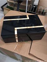Black lacquer humidor trinket box with cream colored inlay in geometric angular pattern on top and side, placed on cardboard box.