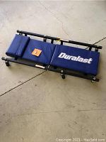 Full view of Duralast mechanics creeper showing padded blue surface and caster wheels on concrete floor.