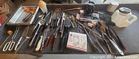 Full overview of kitchen utensils laid on a table including knives, spatulas, scissors, trays, and other miscellaneous tools.