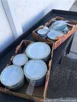 Noritake china dish set arranged in cardboard boxes, showing multiple stacks of dinner and salad plates with green and white floral trim along the edges.