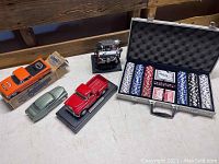 Photo showing the professional poker chip set in a metal case with chips, cards, and dealer button, alongside four diecast model vehicles including 1955 Chevy 3100 red stepside truck, green Mercury car, Chevy small block engine model, and boxed orange Chevy El Camino