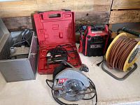 Lot of power tools showing five main items on a table against a wood backdrop, including metal boxed sander, red plastic boxed jigsaw, mobile power pack, extension cord reel, and a Skil circular saw