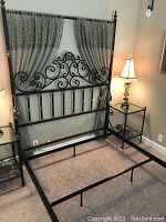 Queen iron bed frame with scrollwork headboard and footboard, flanked by two matching iron nightstands with glass tops and two table lamps.