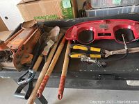 Photo showing assortment of hand tools including car jack, shovel, pickaxe, pry bar, pneumatic sander, kneepads, and garden tools.