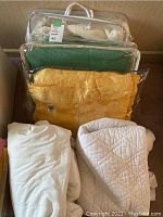 Photo showing three blankets and two mattress protectors stacked, some in clear plastic storage bags. Blankets include green and yellow wool, plus white quilted. Mattress protectors are white and quilted.
