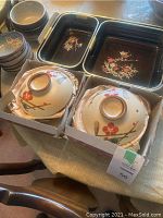 Two ceramic serving bowls with lids and six small floral patterned bowls around them, along with two rectangular nesting trays in background.