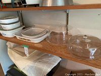 Photo showing four Corning Ware casserole dishes stacked with lids on the left and two clear cake plates with dome lids on the right on wooden shelving.