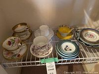 Overview of 7 assorted bone china cups and saucers with additional plates and dishes on white wire shelving. Various floral and gold trim designs.