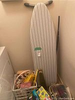 Full view of ironing board with striped cover leaning against the wall, laundry baskets and supplies in front