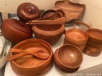 Display of two large wooden salad bowls with lids, several smaller wooden bowls, and wooden salad utensils on top of one large bowl.
